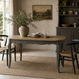 A rustic dining table with a light oak top and dark grey carved legs, styled in a cosy room with chairs, a woven rug, and a large vase of greenery on top.
