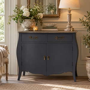 A dark grey wooden cabinet with curved legs, two drawers, and two doors, styled in a bright living room setting with a lamp, flowers, and a mirror.