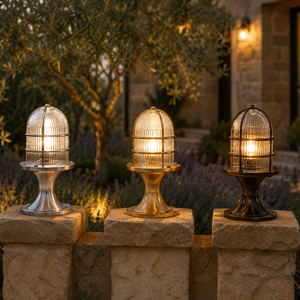 Three outdoor birdcage-style post lights in silver, gold, and bronze finishes mounted on stone pillars in a garden setting at dusk.