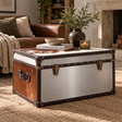 A vintage-style trunk coffee table with a brushed silver metal top and brown leather trim, styled in a cozy living room setting.