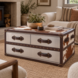 A rustic-style storage trunk coffee table with cowhide-patterned panels and dark brown leather handles, styled in a cosy living room setting.
