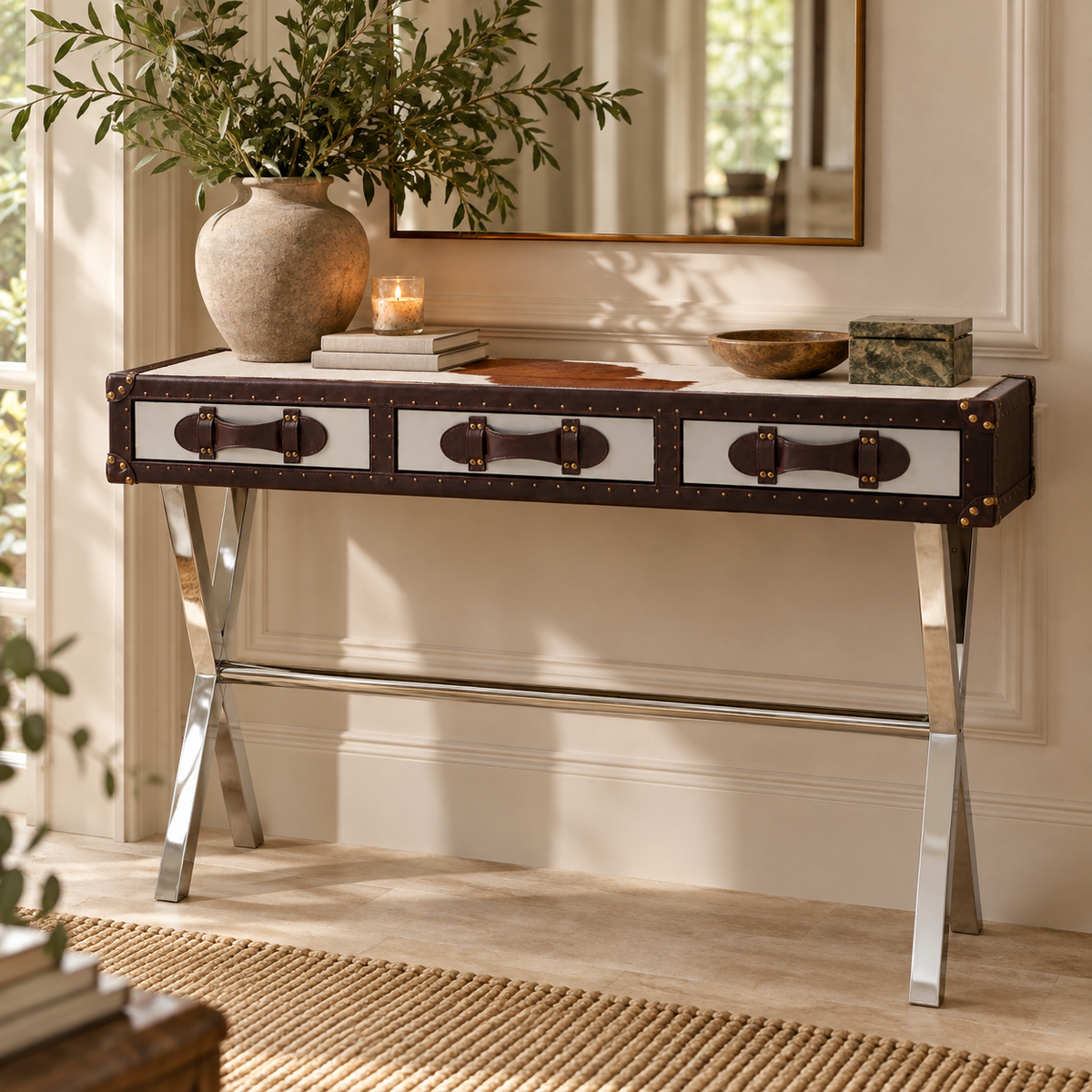 A console table styled in a bright room, featuring a dark brown and white trunk-inspired top with metal handles and legs, styled with a large vase of greenery, books, and decorative bowls.