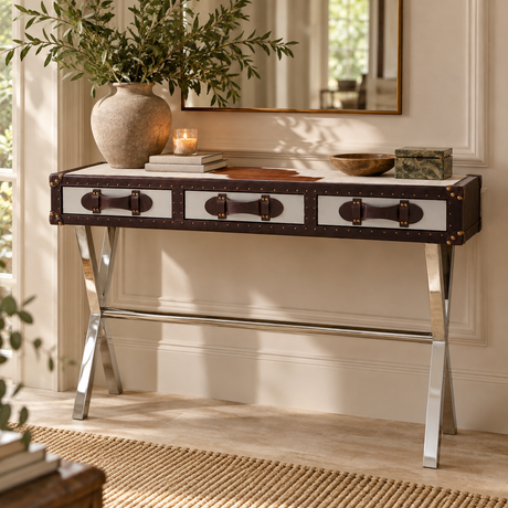 A console table styled in a bright room, featuring a dark brown and white trunk-inspired top with metal handles and legs, styled with a large vase of greenery, books, and decorative bowls.