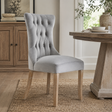A light grey velvet dining chair with a high button-tufted back and light wood legs, styled in a dining room next to a rustic wooden table.
