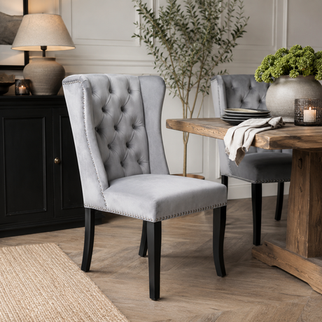 A light grey tufted velvet dining chair with black legs and nailhead trim, styled in a dining room next to a wooden table.