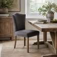 A dark grey upholstered dining chair with nailhead trim and wooden legs, shown in a styled dining room next to a rustic wooden table.