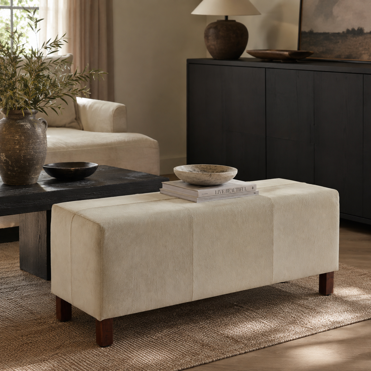 A rectangular upholstered ottoman bench in a warm cream fabric with dark wooden legs, styled in a modern living room setting on a woven rug.