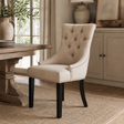 A beige upholstered dining chair with button tufting, nailhead trim, and black legs, shown in a styled dining room setting next to a wooden table.