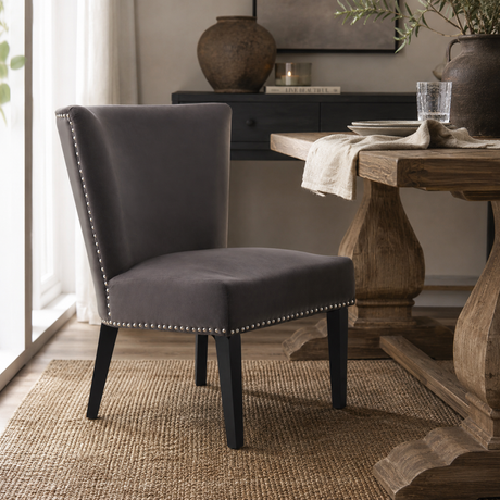 A dark grey upholstered accent chair with black legs and silver nailhead trim, positioned in a dining room next to a rustic wooden table.