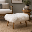 A round white sheepskin ottoman stool with four light wood legs, styled in a cosy living room setting on a woven rug next to a wooden armchair and side table.