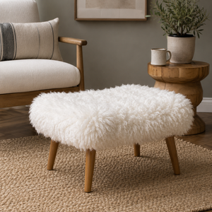 A round white sheepskin ottoman stool with four light wood legs, styled in a cosy living room setting on a woven rug next to a wooden armchair and side table.