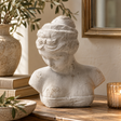 Woman's Head Icon bust on a wooden sideboard beside a stack of books, a stoneware vase of olive branches and a lit candle in a glass holder