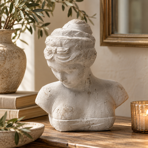 Woman's Head Icon bust on a wooden sideboard beside a stack of books, a stoneware vase of olive branches and a lit candle in a glass holder