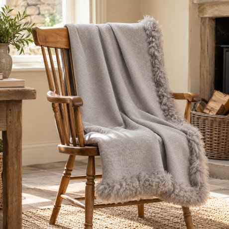 Woven Grey Wool Blanket With Sheepskin Trim draped over a wooden Windsor chair by a stone fireplace with a log basket and olive branches