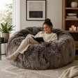 Woman reading a book on a fluffy grey sheepskin bean bag chair in a cosy living room.
