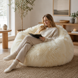 Woman reading a book on a fluffy white sheepskin bean bag chair in a cosy living room.