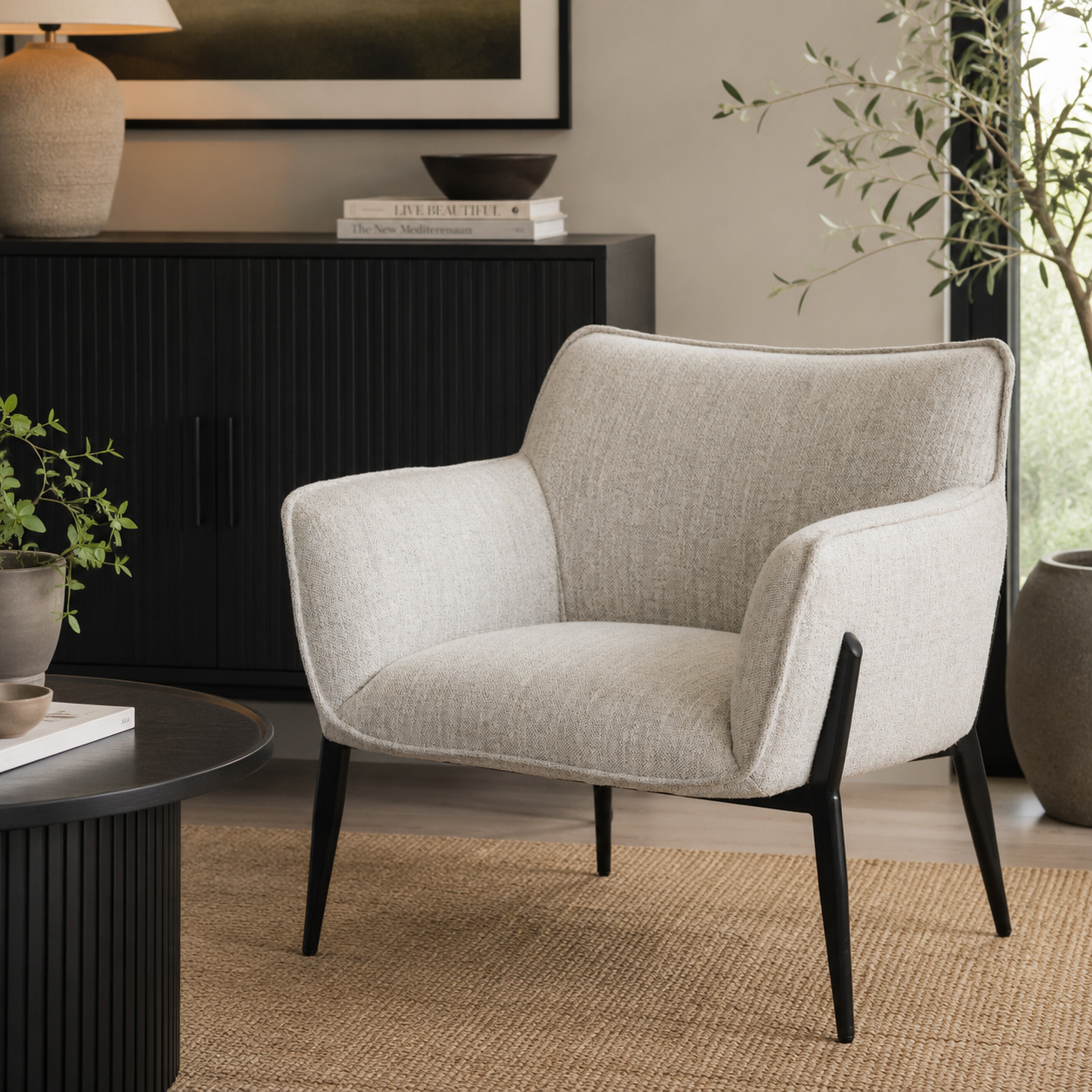 A light beige textured fabric armchair with black tapered legs, styled in a modern living room setting next to a black sideboard and a round black coffee table.