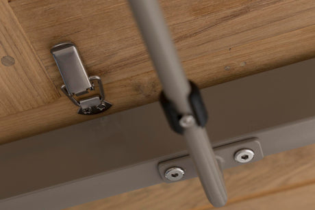 Close-up of a hinge with a brushed silver metal latch mechanism attached to a light wood grain table, with a grey metal support rail and bracket below.