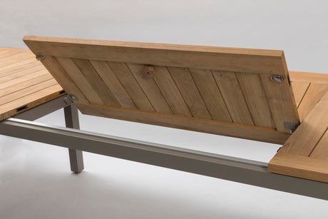 Close-up of a wooden outdoor lounge table with a butterfly leaf extension and light gray metal frame, photographed against a plain light grey studio background.