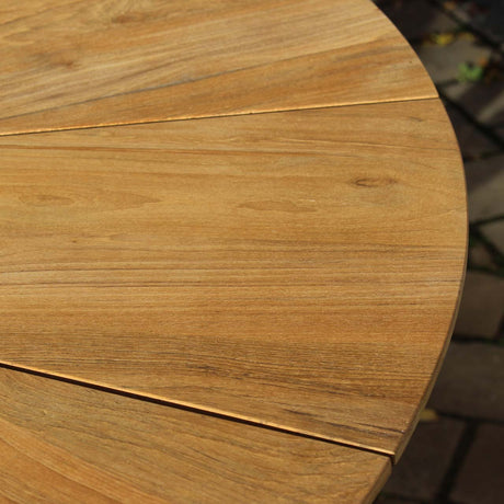 Close-up of a round wooden table with a warm medium-brown finish and visible wood grain, with patio paving in the background.