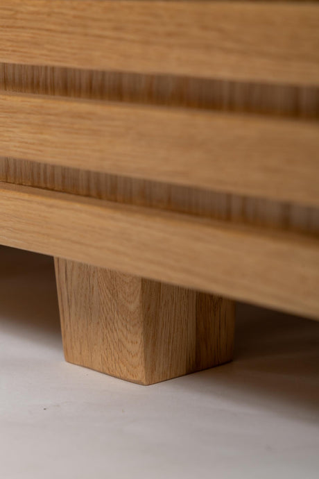 Close-up of a light oak wood furniture piece showing the wood grain, horizontal grooved details, and a block-style leg on a neutral floor.