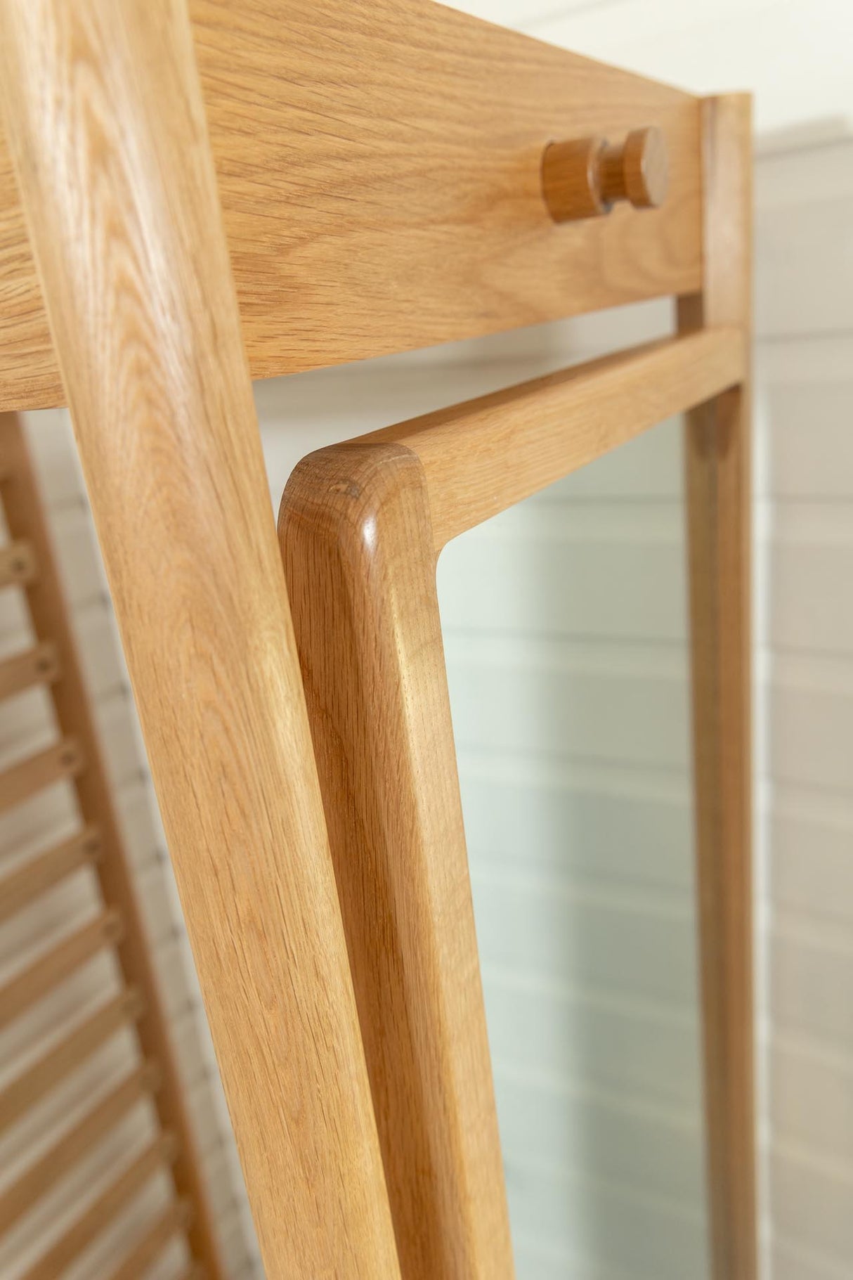 Close-up of a light oak wood frame with rounded corners and a small round wooden knob, showing part of a mirror and a white paneled wall in the background.