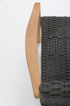 Close-up detail of a chair with a curved light natural wood armrest and a dark charcoal grey woven seat cushion on a white background.