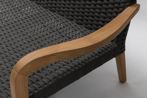 Close-up of a woven outdoor sofa showing dark charcoal grey rope upholstery, a curved natural wood armrest, and light wood legs on a light grey floor.