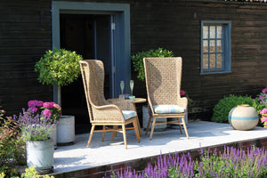 Two tall woven wicker patio chairs with light wood legs and striped cushions sit on a stone patio in front of a dark wood house, with potted plants and flowers around them.