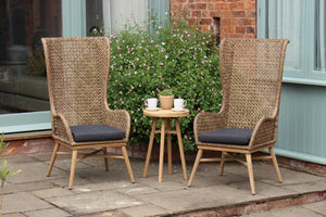 Two tall woven rattan patio chairs with dark gray seat cushions and a small round wooden side table between them, set on a stone patio in front of a house with greenery and a brick wall.