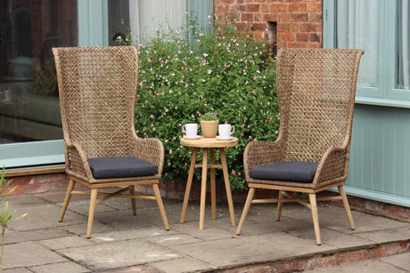 Two tall woven rattan patio chairs with dark gray seat cushions and a small round wooden side table between them, set on a stone patio in front of a house with greenery and a brick wall.