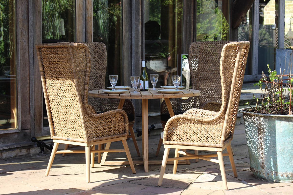 Three woven rattan outdoor dining chairs with light wood frames arranged around a small round wooden table on a patio, with wine glasses and bottles on the tabletop.