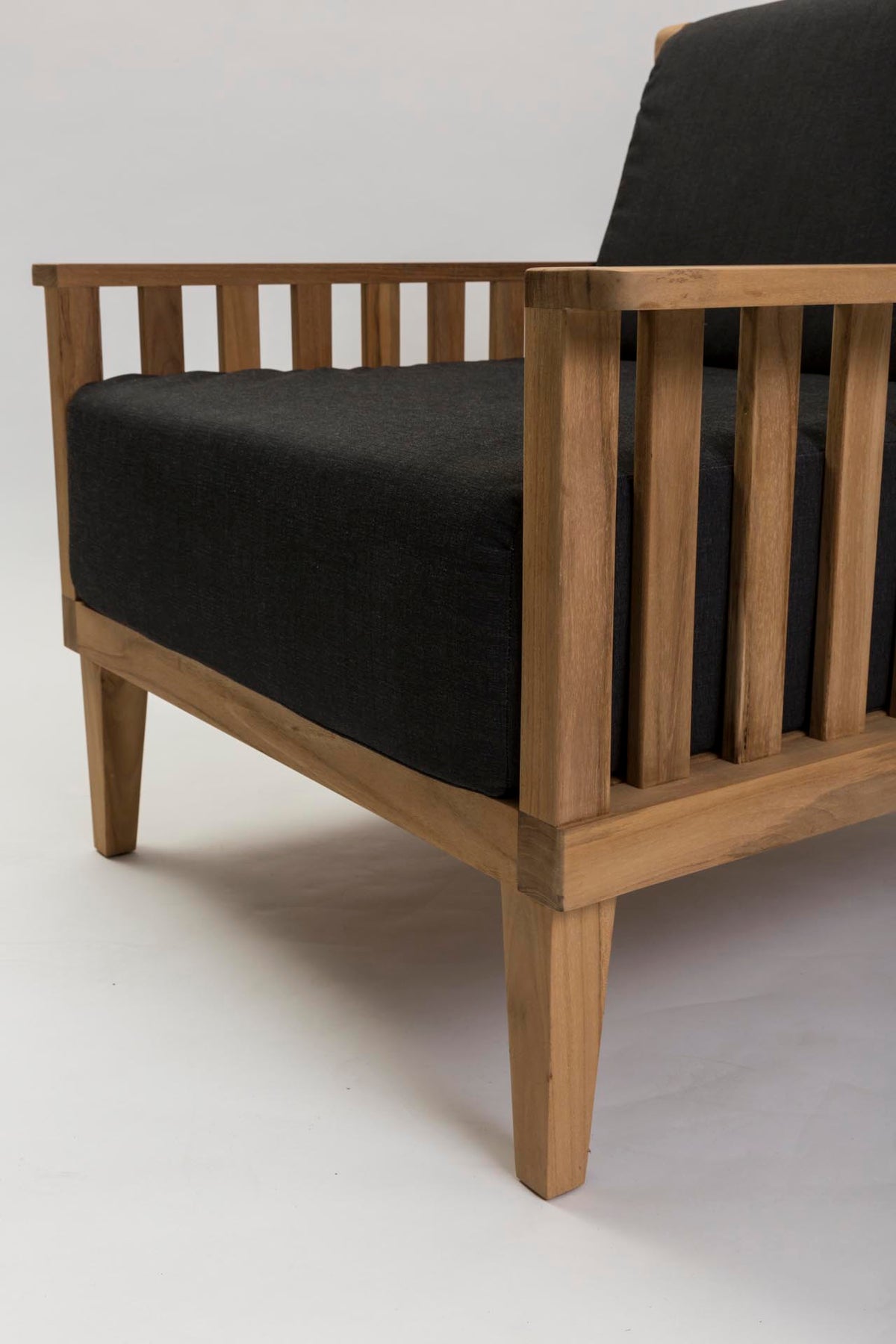 A close-up of a wooden armchair with a natural wood frame and thick black fabric cushions, shown against a plain light gray studio background.