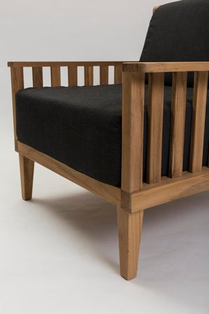 A close-up of a wooden armchair with a natural wood frame and thick black fabric cushions, shown against a plain light gray studio background.
