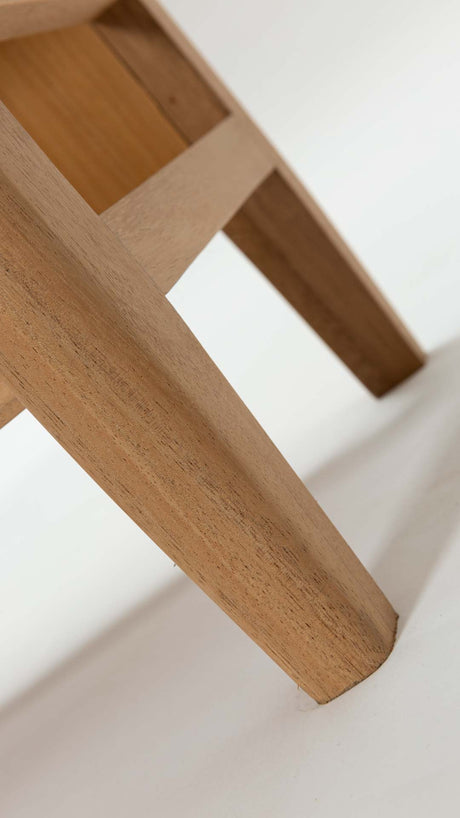 Close-up of a light natural wood stool leg and crossbar on a white floor against a white wall.