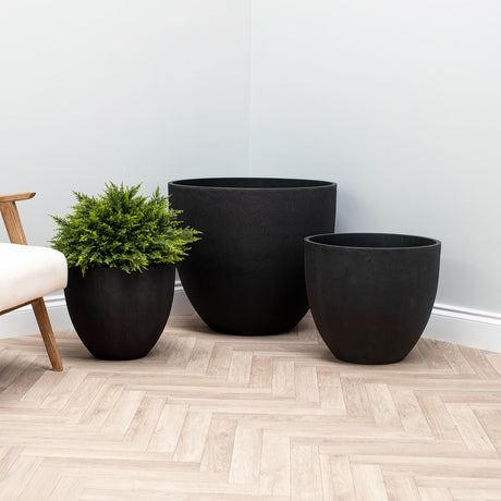 Three matte black planters in different sizes arranged on a light wood herringbone floor against a pale gray wall, with a small green plant in the left planter and part of a chair visible at the left edge.