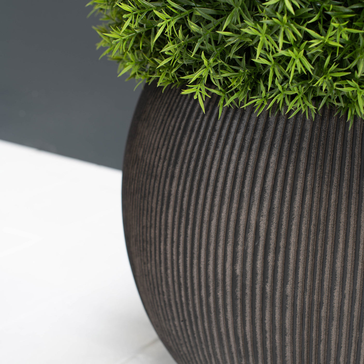 A close-up of a ribbed dark brown planter pot filled with bright green artificial foliage, set against a split white and dark grey background.