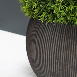 A close-up of a ribbed dark brown planter pot filled with bright green artificial foliage, set against a split white and dark grey background.