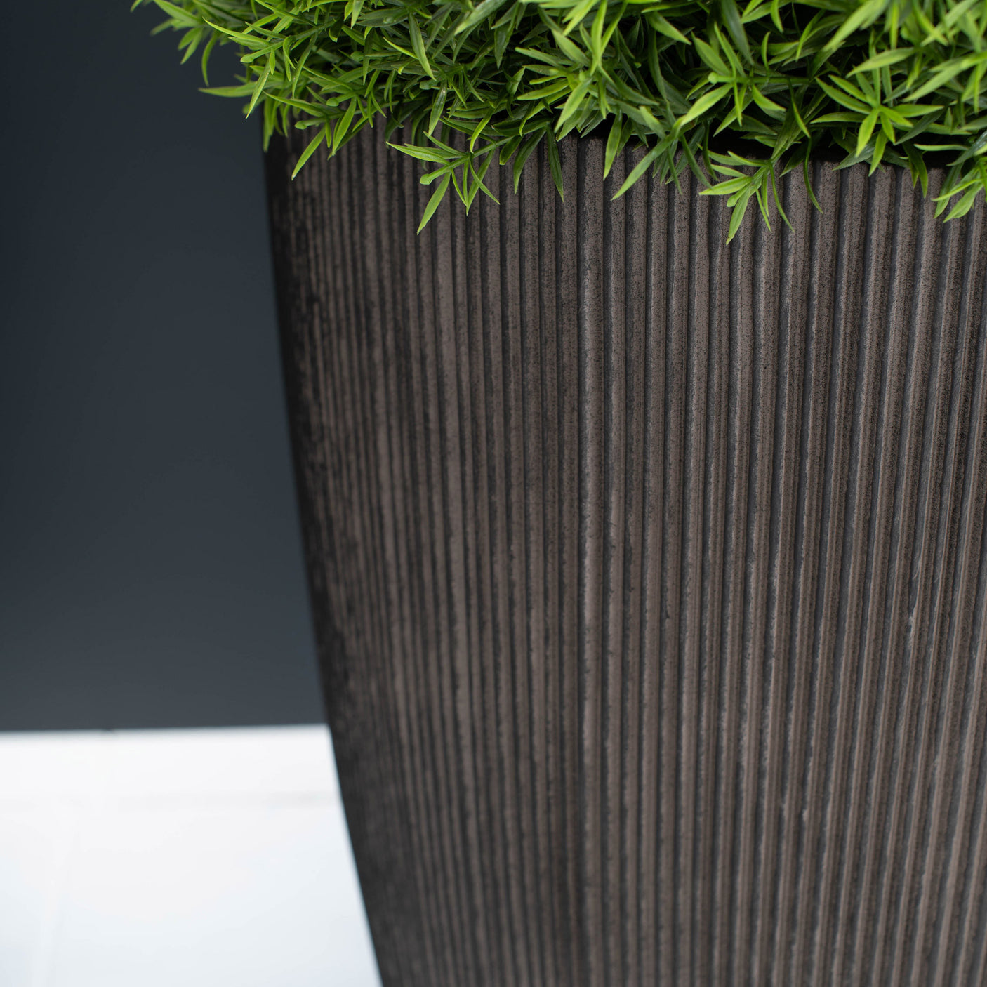 A close-up of a tall, dark brown ribbed planter pot with green foliage emerging from the top, set against a dark grey background and a white surface.