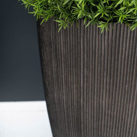 A close-up of a tall, dark brown ribbed planter pot with green foliage emerging from the top, set against a dark grey background and a white surface.