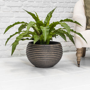 A round ribbed brown planter with a green plant on a light grey floor next to a chair in front of a white brick wall