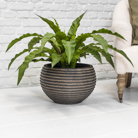 A round ribbed brown planter with a green plant on a light grey floor next to a chair in front of a white brick wall