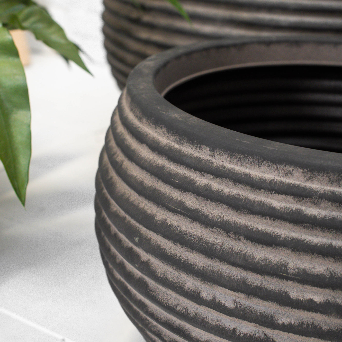 A close-up of a large ribbed planter pot in dark brown, with another similar pot blurred in the background and a green plant leaf entering from the left.
