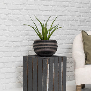 A green aloe vera plant in a dark gray ribbed ceramic pot, displayed on a dark gray wooden slat side table against a white brick wall, with a cream-colored armchair partially visible on the right.