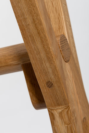 Close-up detail of a wooden chair or stool leg joint, showing natural wood grain, a circular peg, and a dowel on a light gray background.